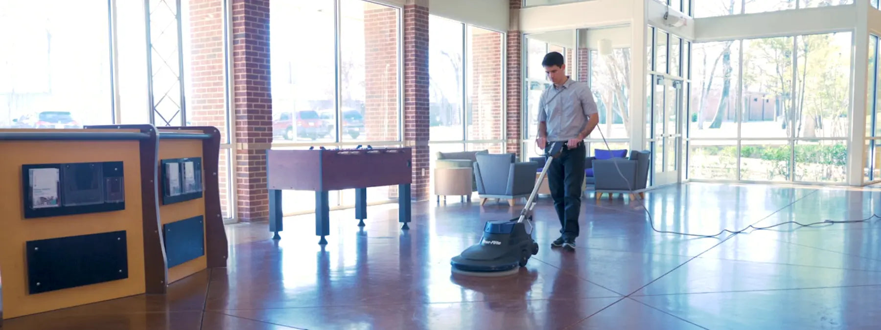 Operator running a Powr-Flite floor burnisher across a glossy, reflective lobby floor beside tall windows