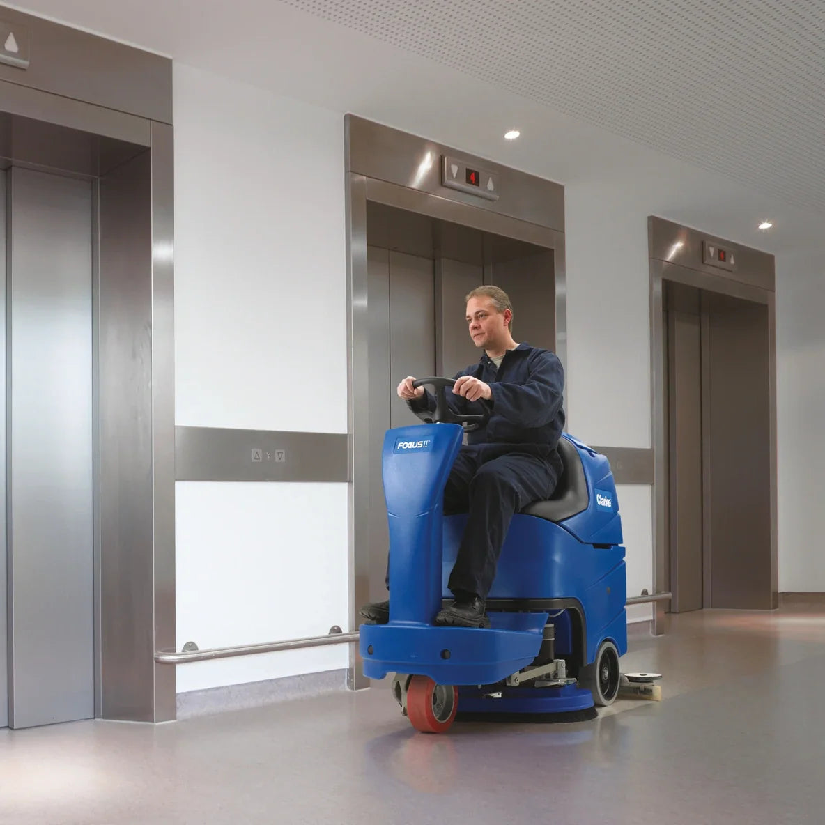 Operator scrubbing an elevator lobby floor with a Clarke Focus II MicroRider, next to the steel elevator doors