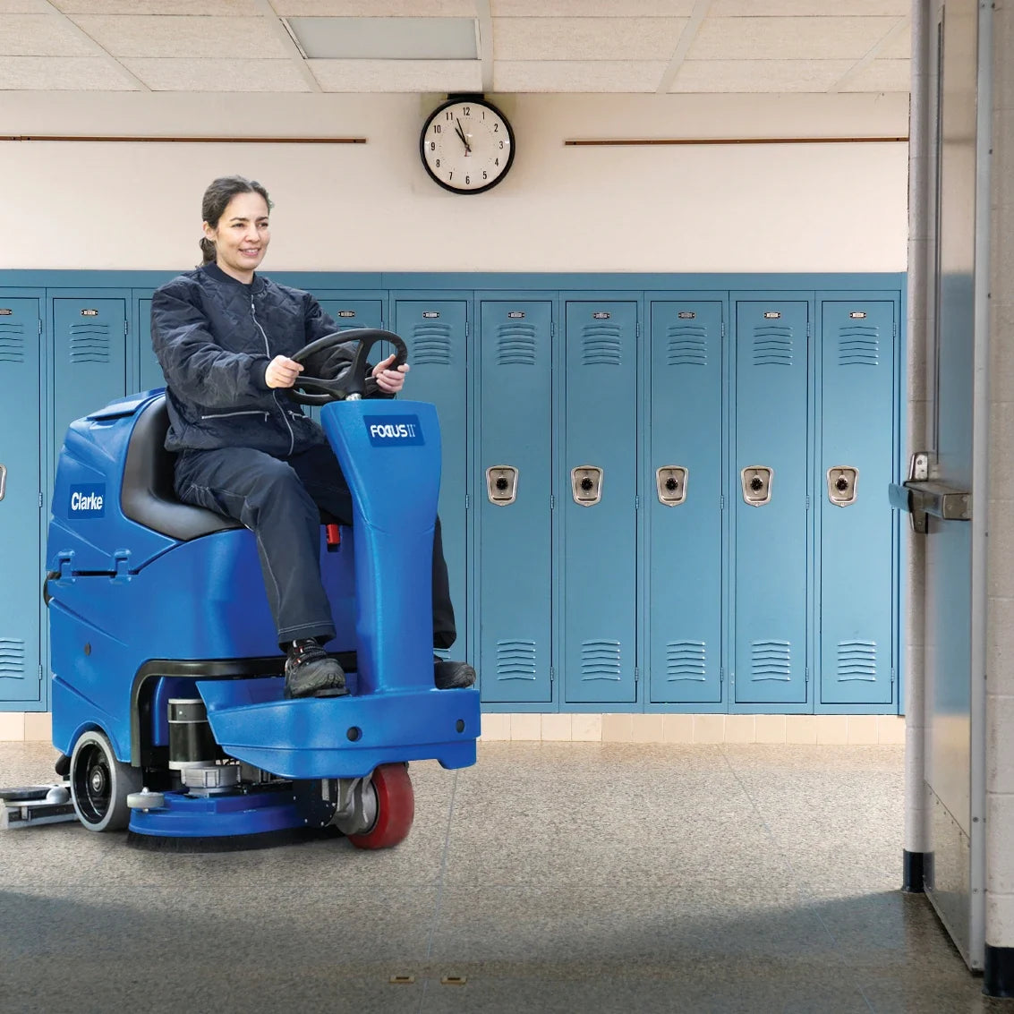 Custodian riding a Clarke Focus II MicroRider through a school hallway lined with lockers, heading through a doorway