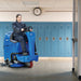 Custodian riding a Clarke Focus II MicroRider through a school hallway lined with lockers, heading through a doorway
