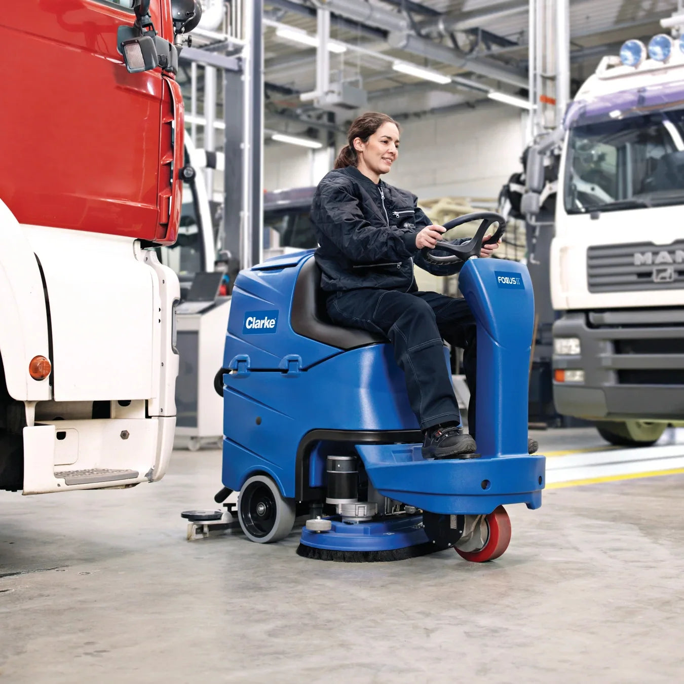 Operator driving a Clarke Focus II MicroRider disc scrubber across the floor of a truck service bay