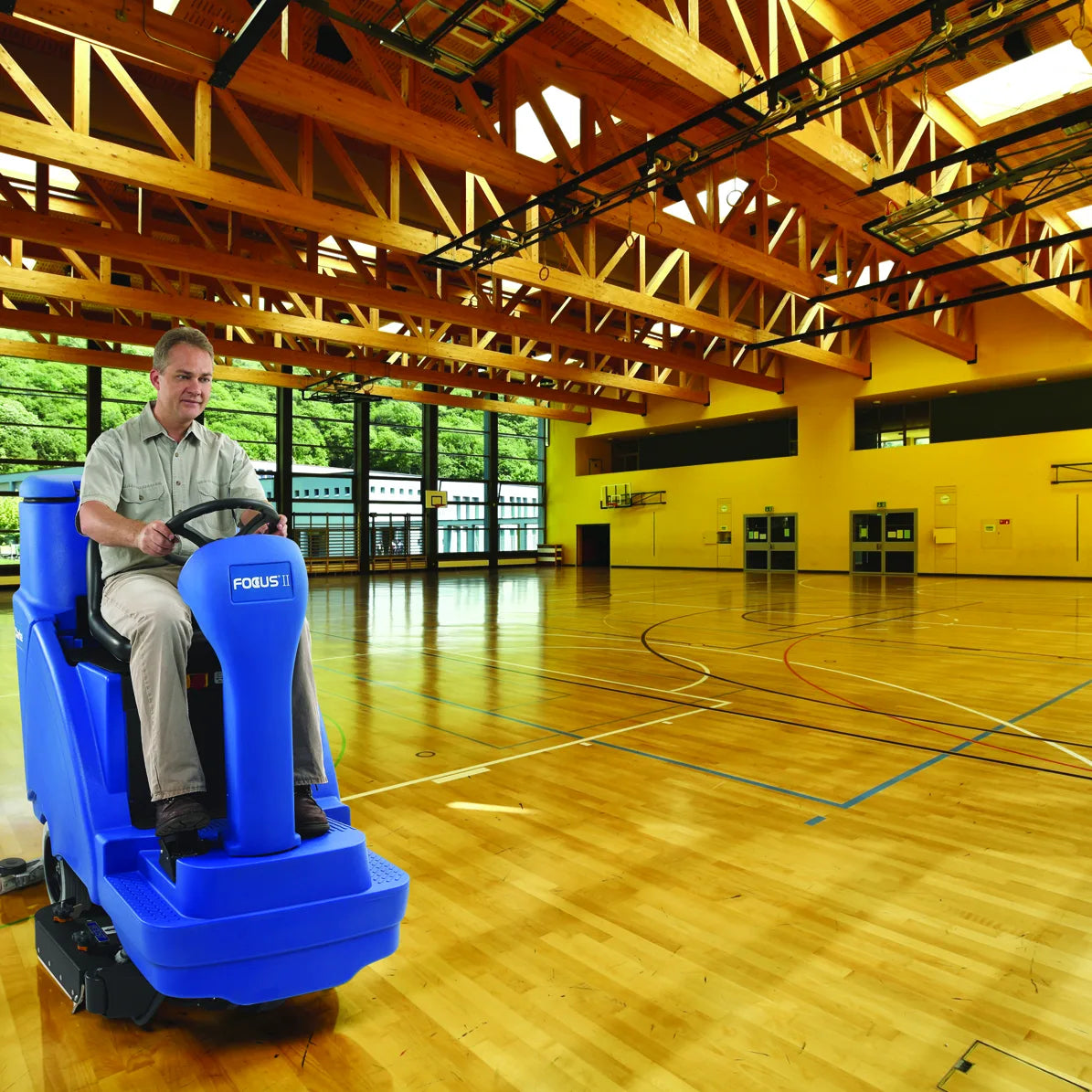 Operator riding a Clarke Focus II Rider across a school gymnasium wood basketball floor