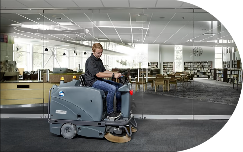 Operator riding the ICE iS1100L+ floor sweeper across an open commercial interior with glass partitions
