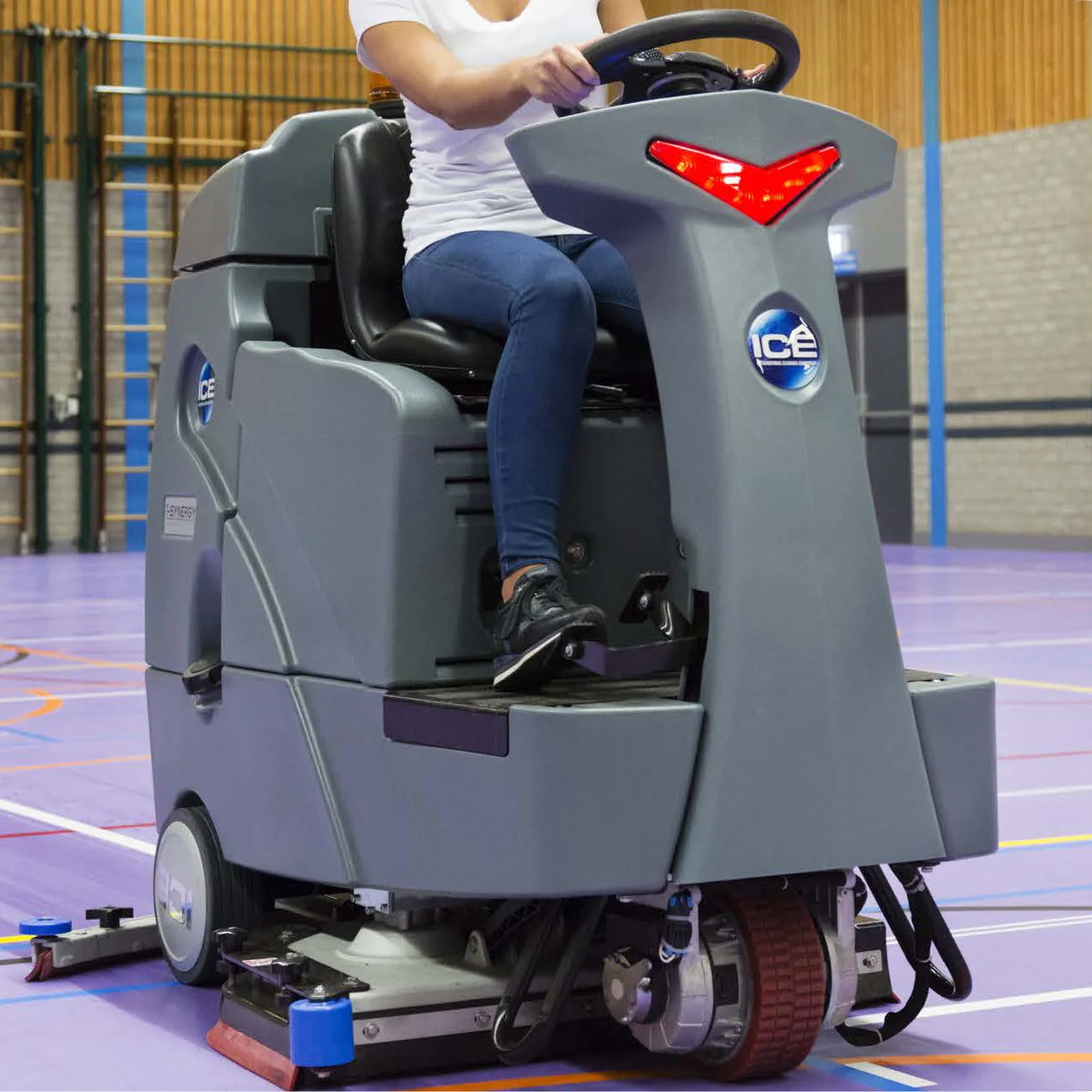 Operator driving the ICE RS26+ ride-on scrubber across a purple sports hall floor, squeegee down and pad drivers running