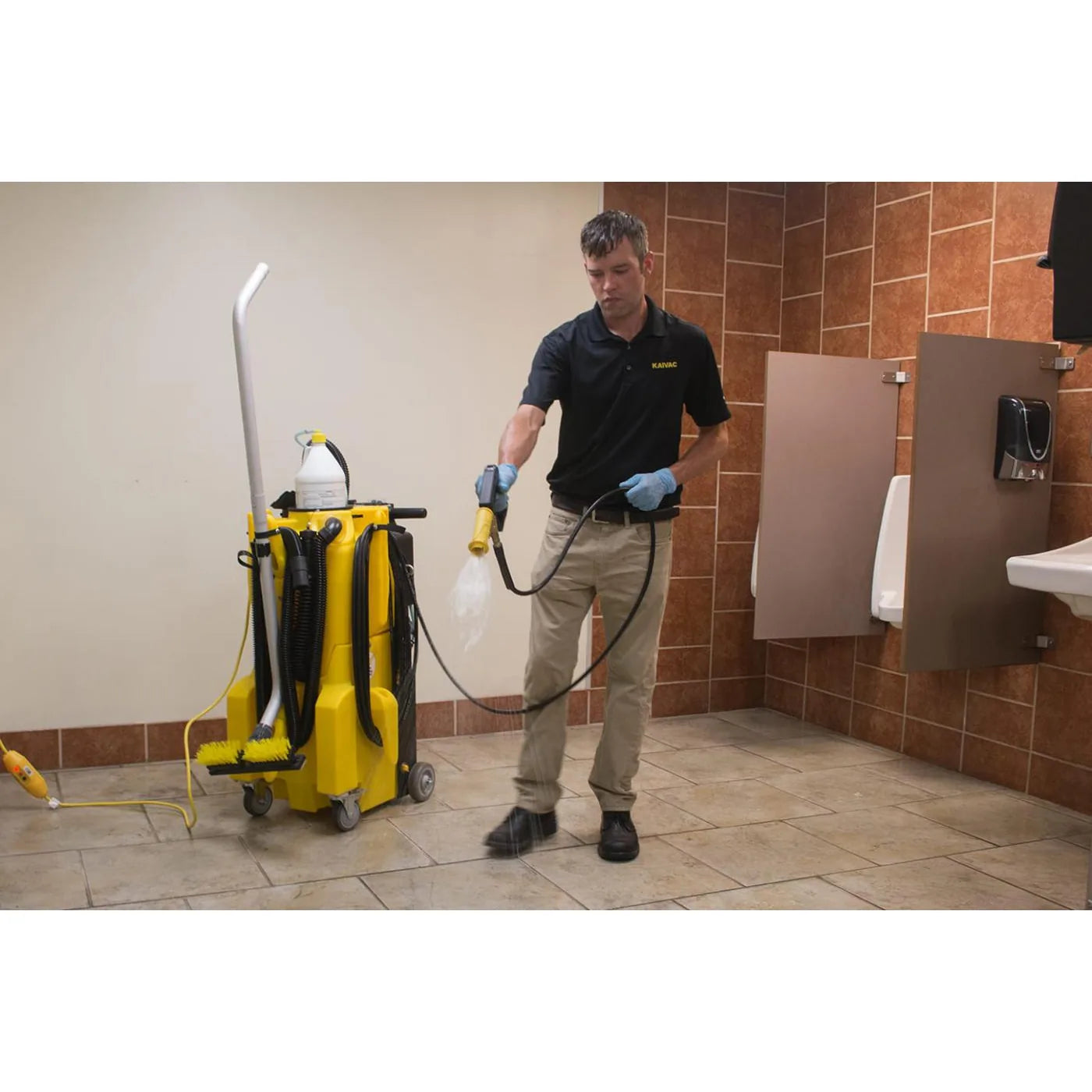 Custodian in gloves rinsing a tiled restroom floor with the KaiVac 1250 spray gun, machine parked beside him