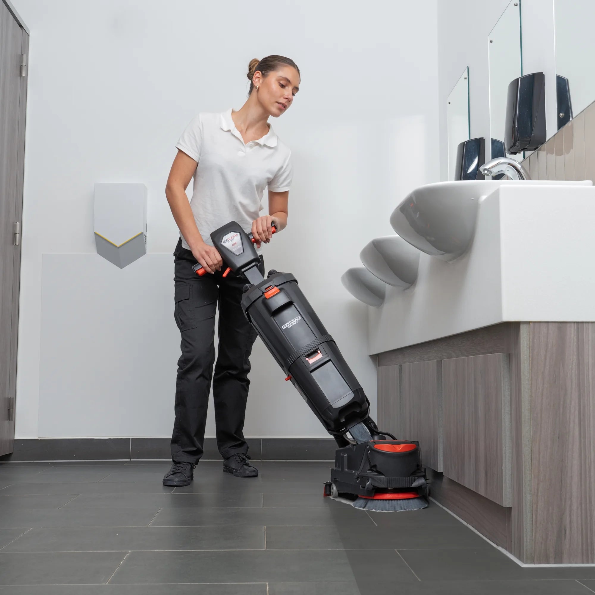 Custodian scrubbing a restroom tile floor under the sink counter with the NaceCare 220NX