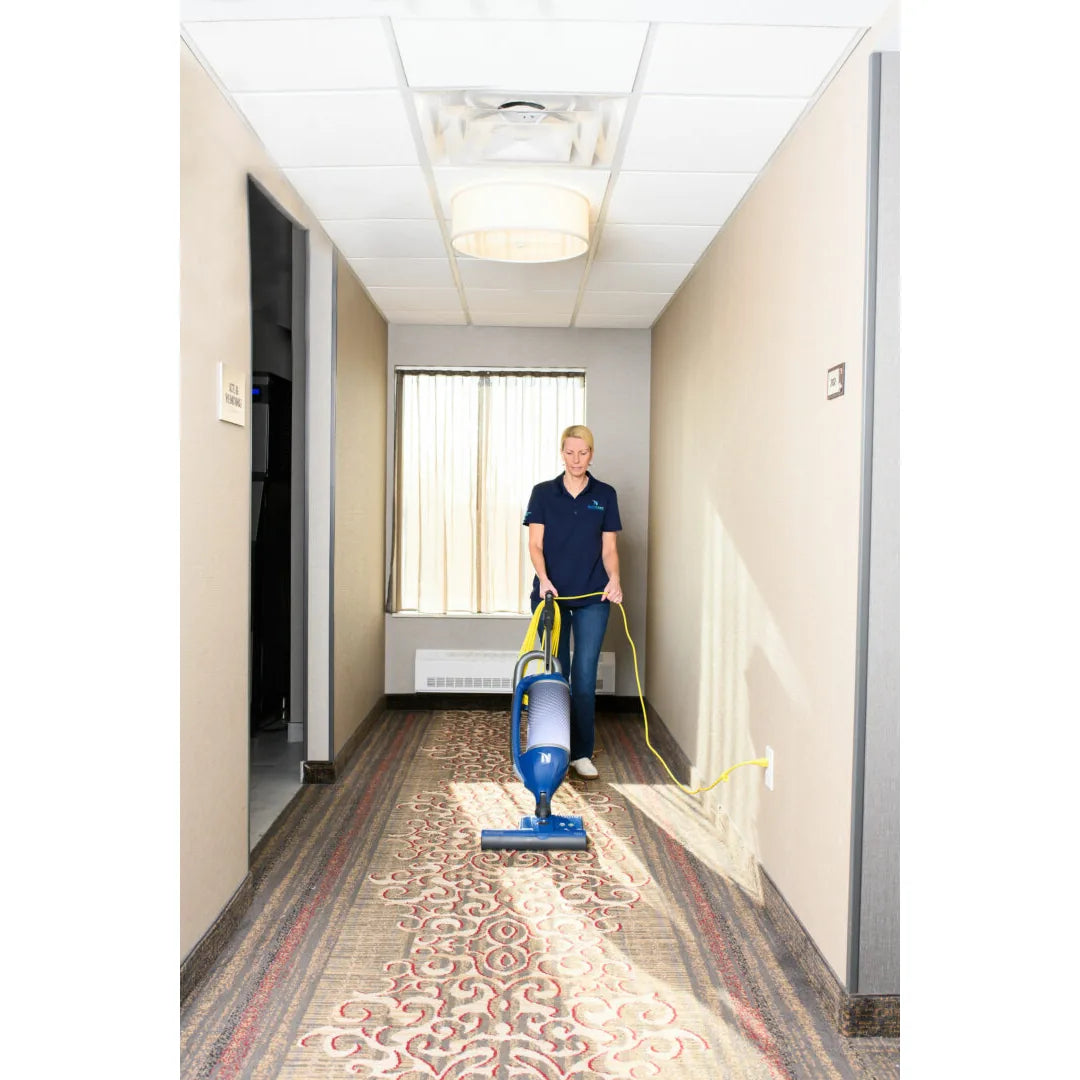 Housekeeper vacuuming a carpeted hotel corridor with a NaceCare upright vacuum