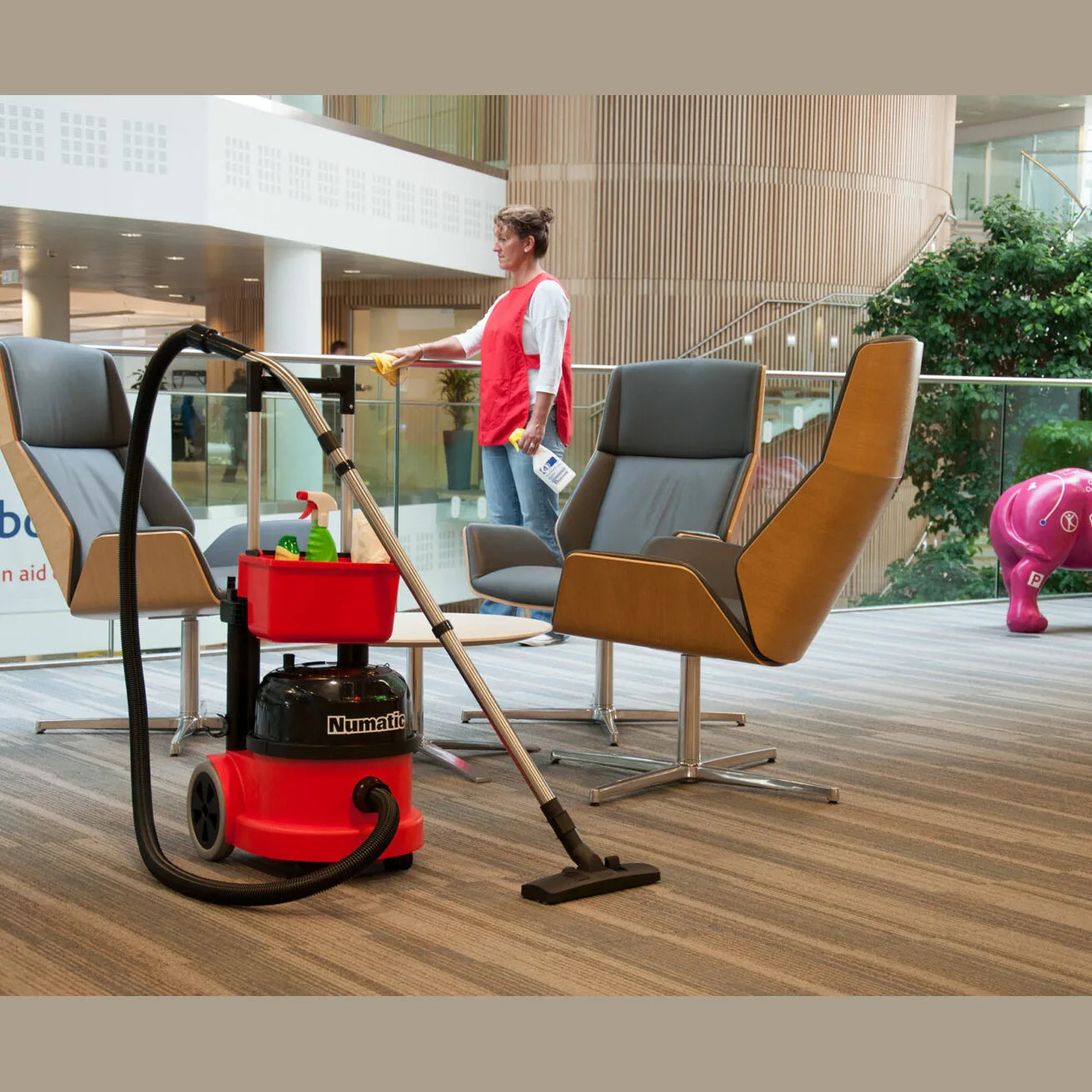 Housekeeper in a red vest wiping down a modern office lounge, the kind of one-person room route the PPR 390 trolley is made for