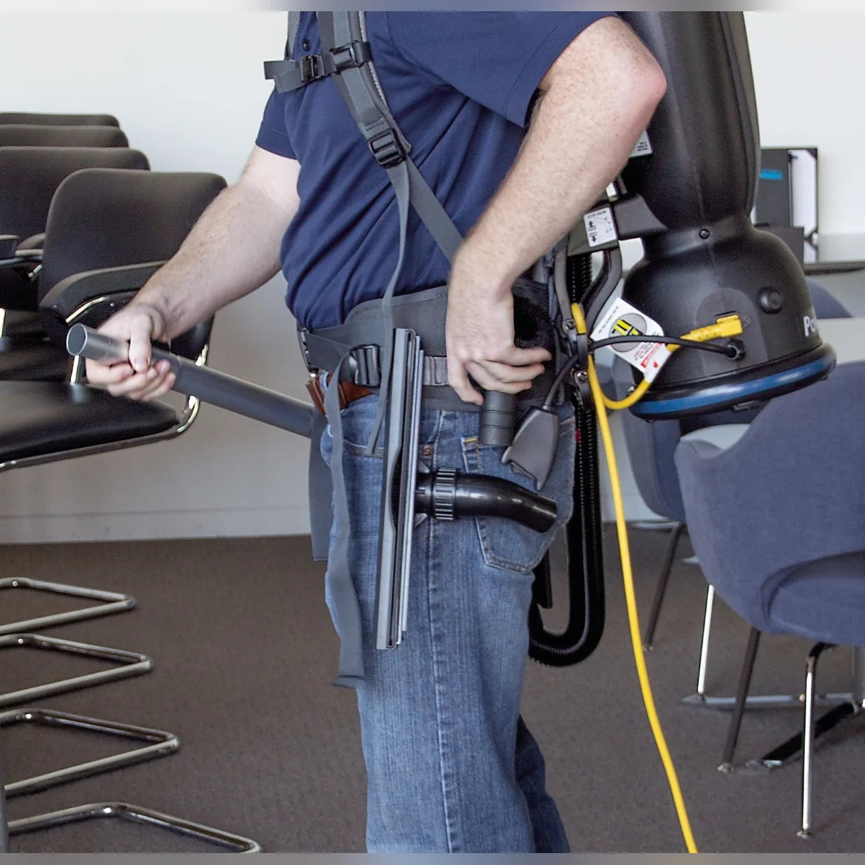 Operator wearing a Comfort Pro backpack vacuum in a meeting room, with tools on the waist belt and the power cord at the hip