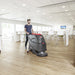 Operator guiding a Viper AS5160 walk-behind floor scrubber across a wood-look floor in a bright canteen