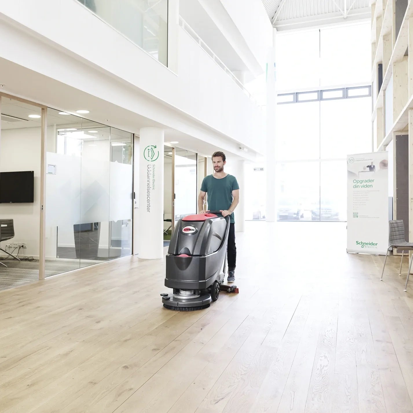 Operator guiding the Viper AS5160T floor scrubber across a light wood-look floor in a bright office atrium